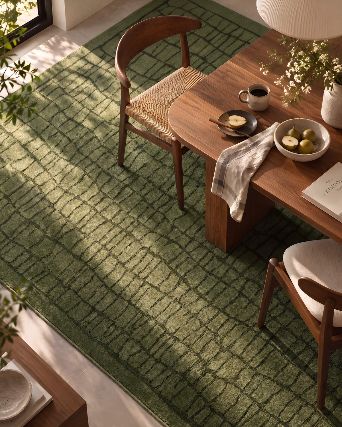 An overhead view of an olive green luxury Wilton jacquard rug highlights its raised grid pattern, rich dimensional texture, and refined contrast beneath a warm wood dining table with natural sunlight and elevated modern decor.