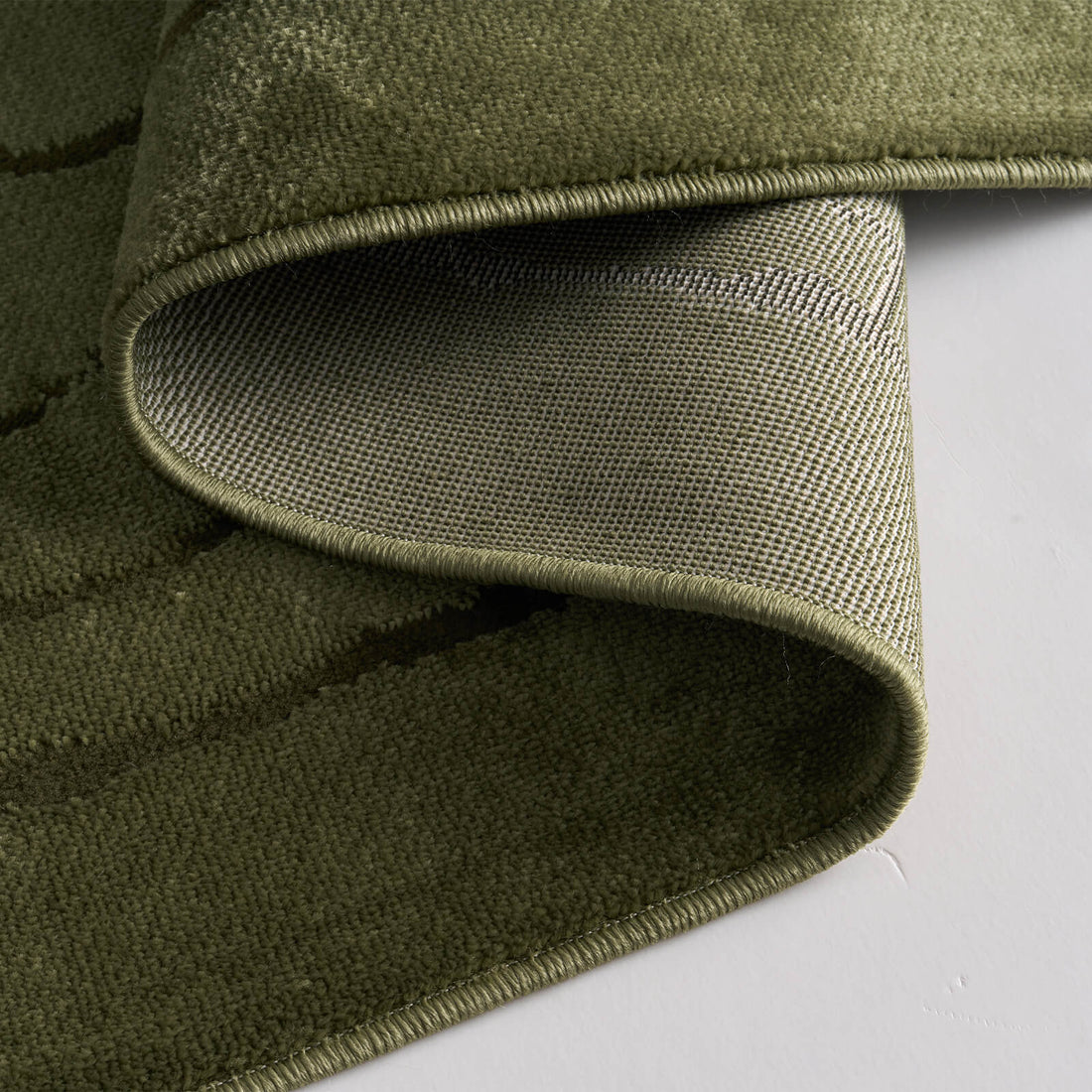 A close-up view of an olive green luxury Wilton jacquard rug showing its curled corner, structured woven backing, plush raised surface, and refined bound edge for elevated living room and bedroom styling.