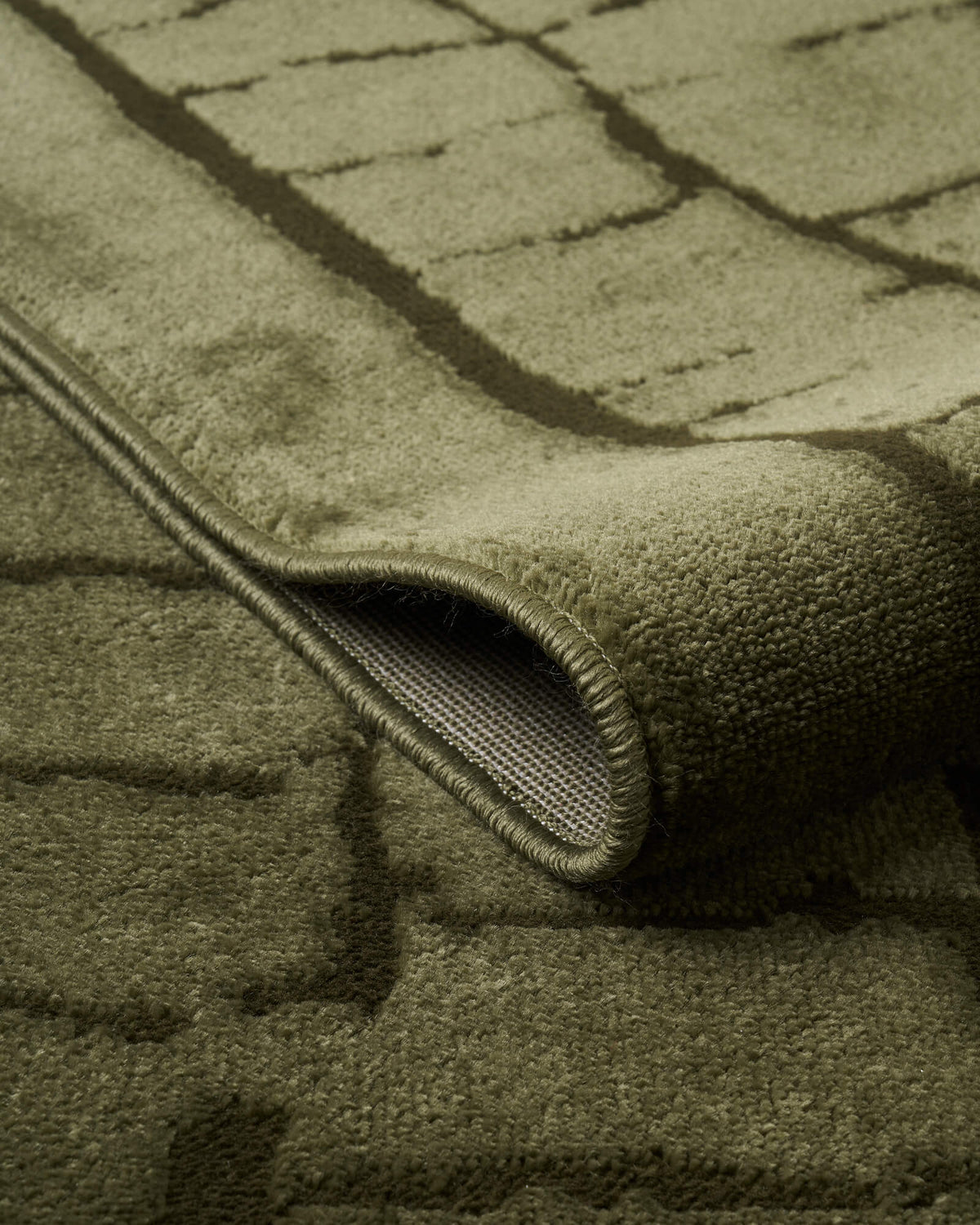 A close-up view of an olive green luxury Wilton jacquard rug showing its folded edge, structured backing, raised grid texture, and refined bound finish for elevated living room and bedroom decor.