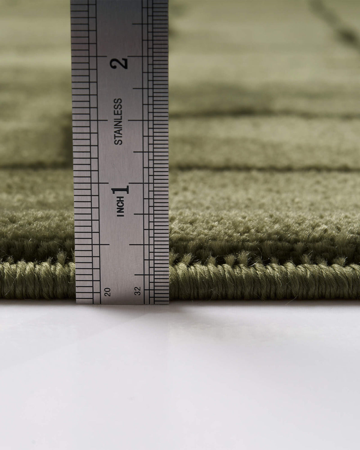 A close-up view of an olive green luxury Wilton jacquard rug showing its ruler-measured pile height, plush raised grid texture, and refined bound edge for elevated living room and bedroom decor.