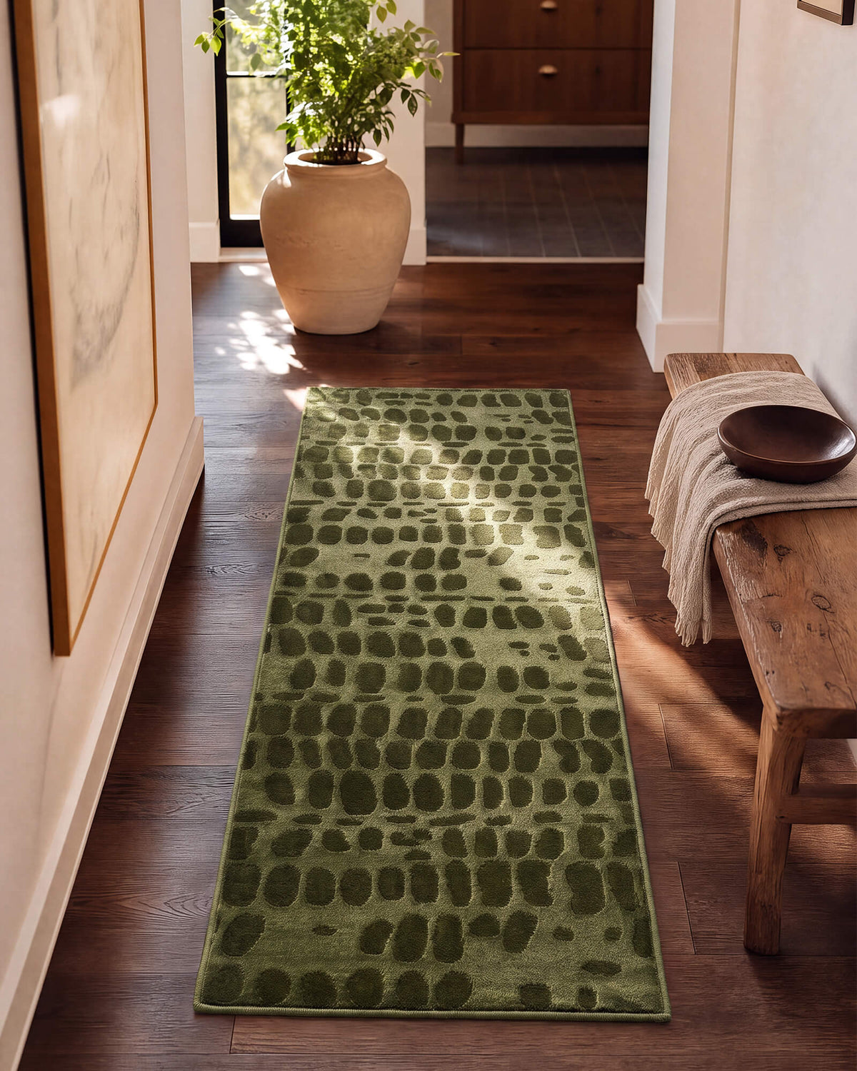 An olive green luxury Wilton jacquard runner rug with raised pebble-inspired texture adds rich sculptural depth to a sunlit entry hallway with warm wood floors, a rustic bench, potted greenery, and refined natural decor.