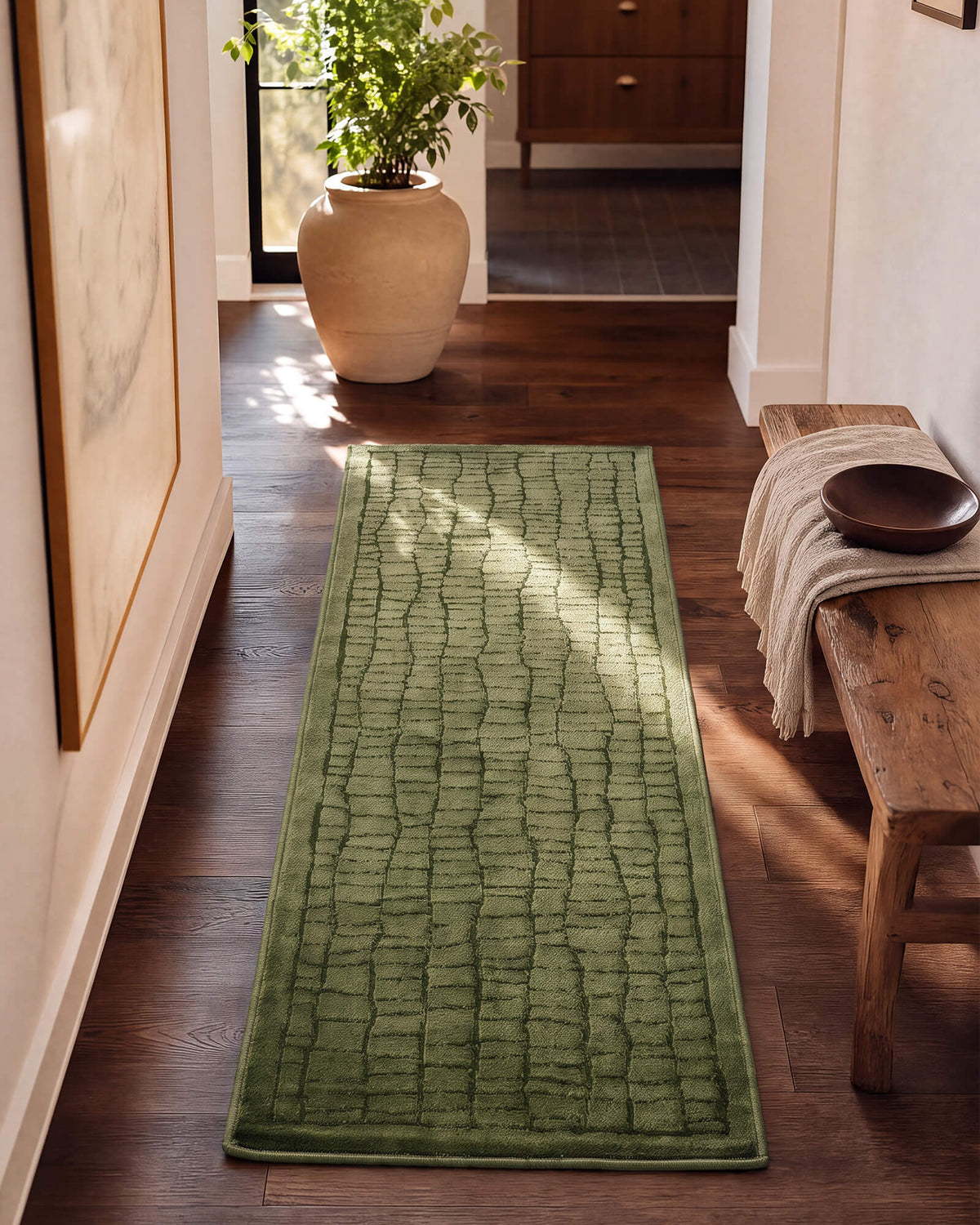 An olive green luxury Wilton jacquard runner rug with a raised crackle texture adds rich tonal depth to a sunlit entryway hall with warm wood floors, a rustic bench, potted greenery, and refined natural decor.