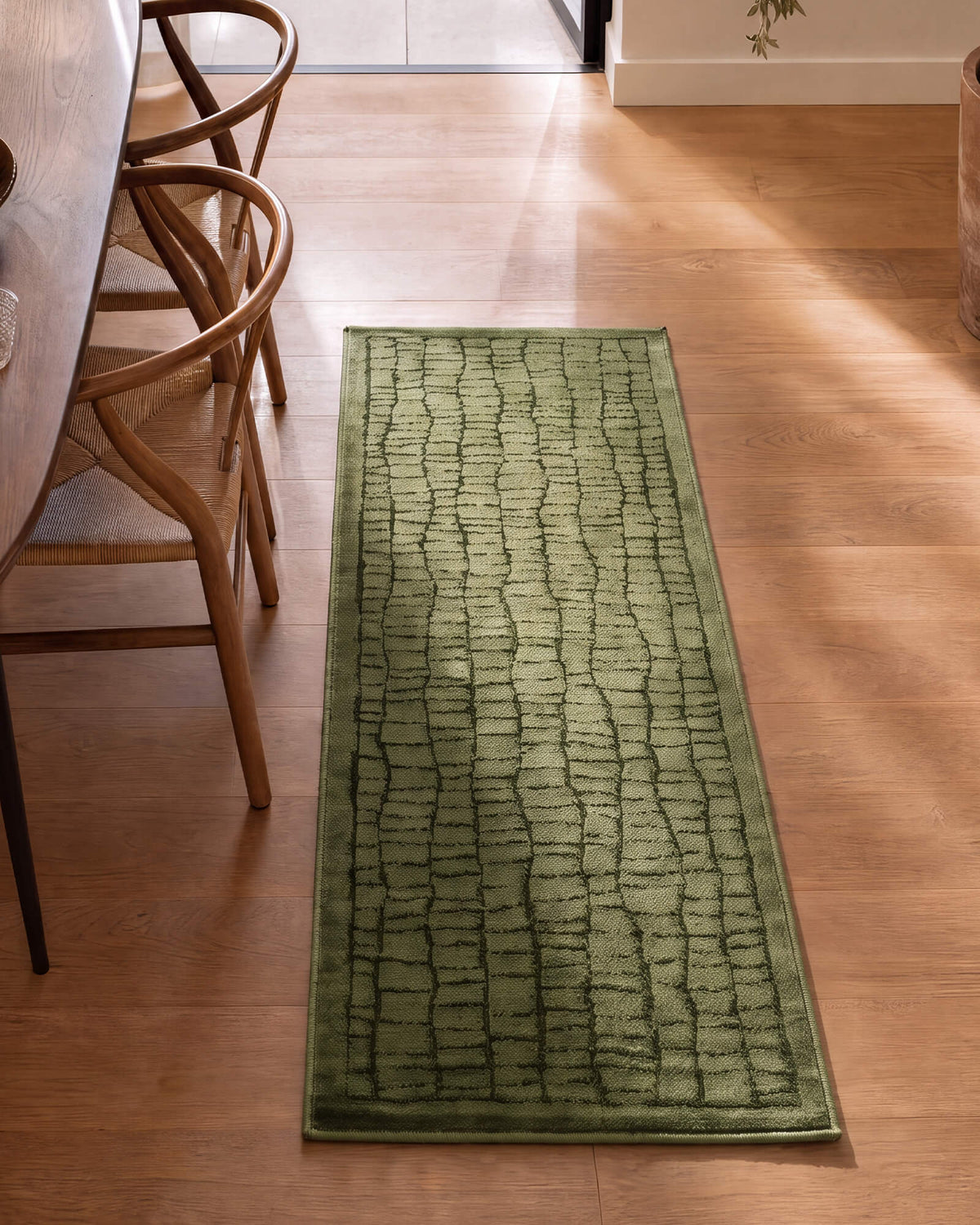 An olive green luxury Wilton jacquard runner rug with a raised crackle grid texture adds rich dimension to a sunlit kitchen hallway with warm wood flooring, woven chairs, and refined modern decor.