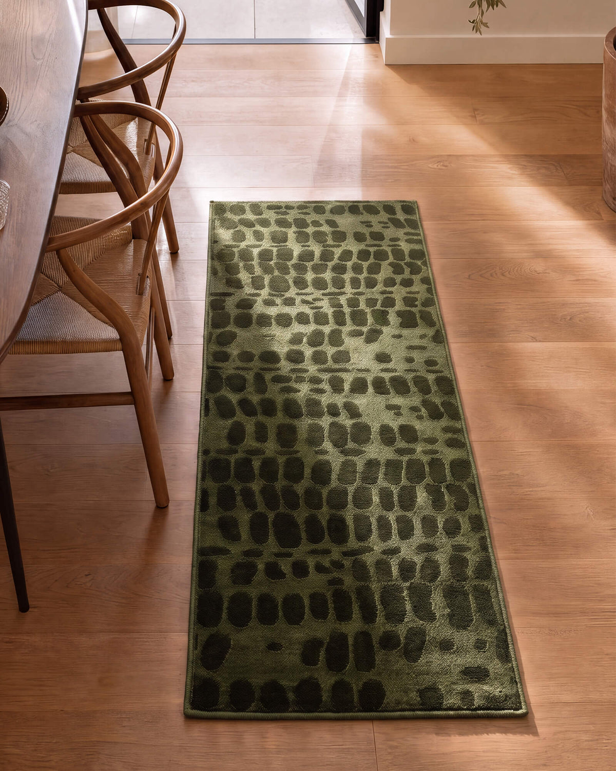 An olive green luxury Wilton jacquard runner rug with a raised pebble-inspired pattern adds rich sculptural texture to a sunlit kitchen hallway with warm wood floors, woven chairs, and refined natural decor.