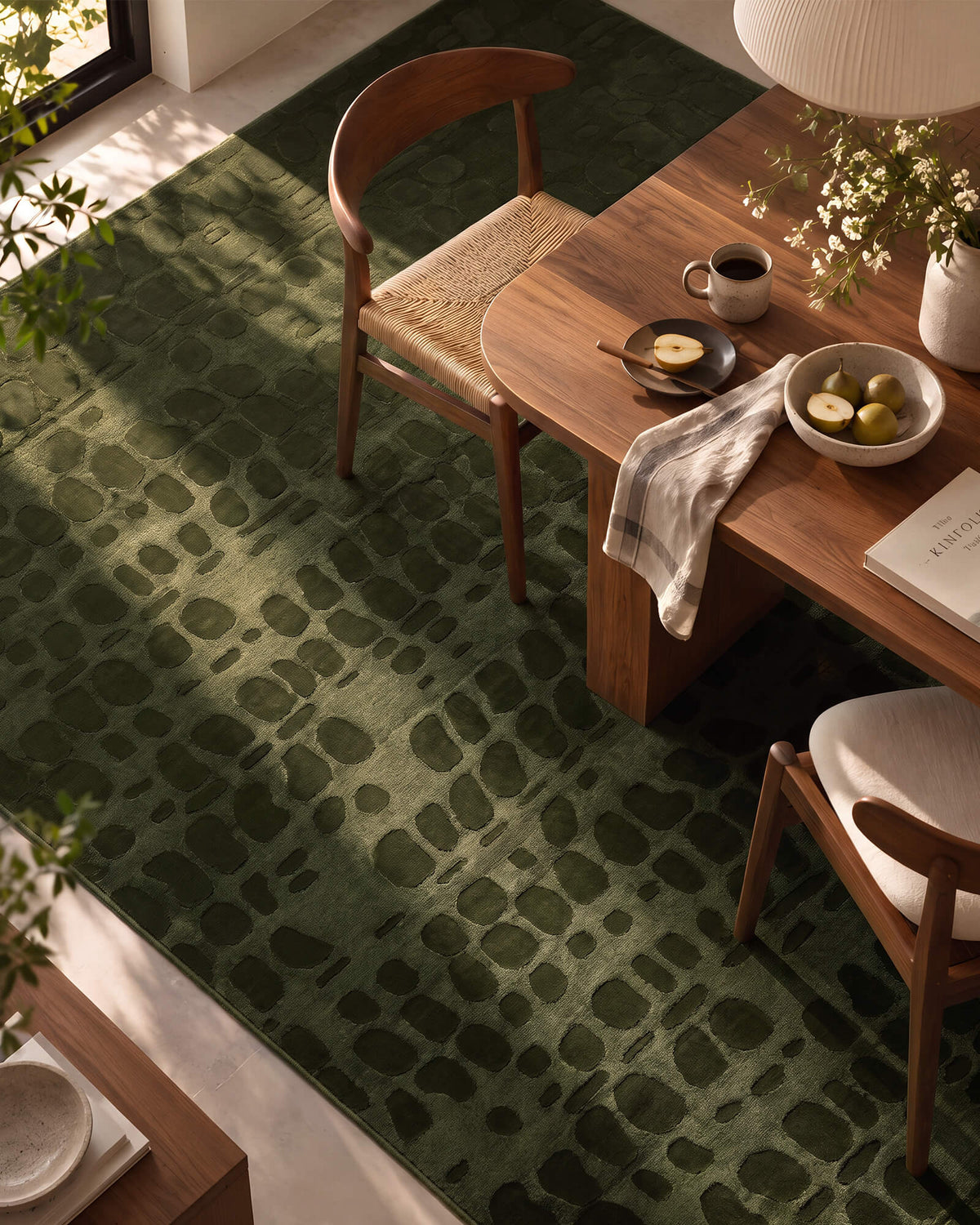 An olive green premium Wilton jacquard rug with raised pebble-inspired texture adds sculptural depth to a refined dining room with warm wood furniture, soft sunlight, and elevated modern decor.