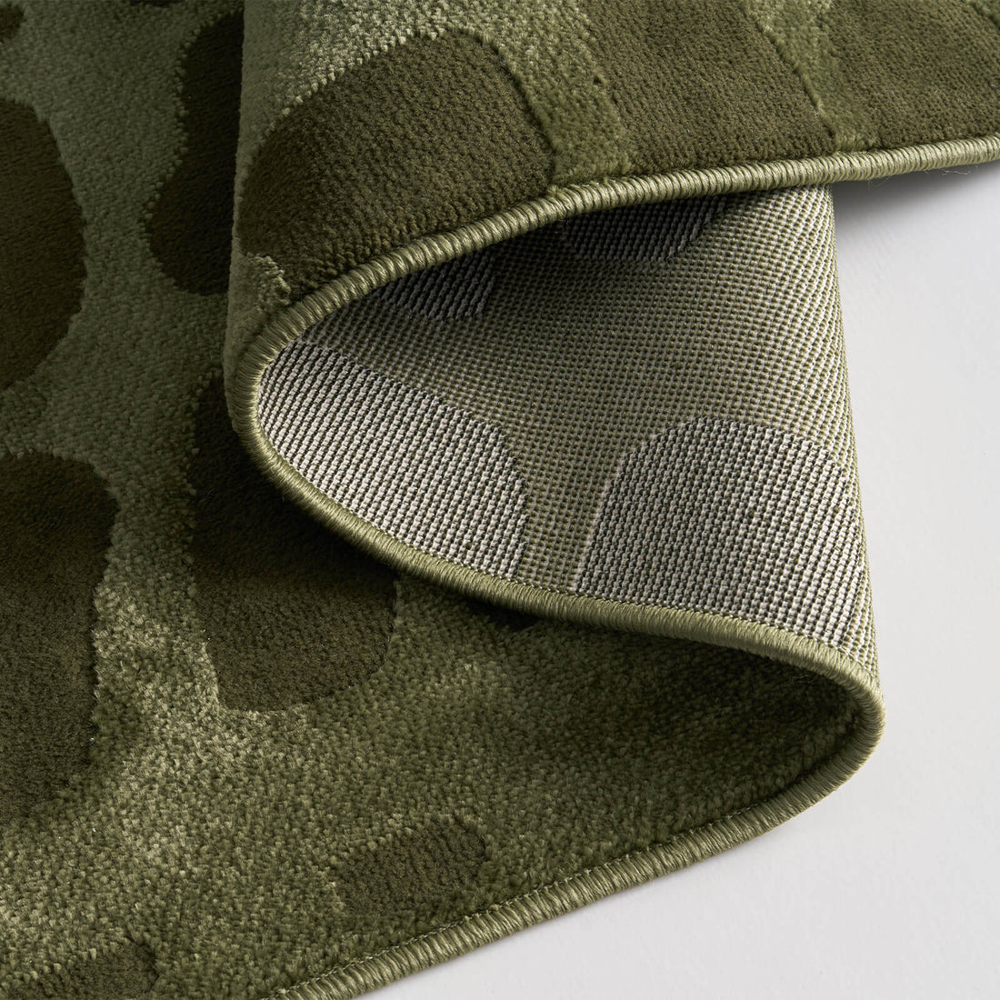 A close-up view of an olive green premium Wilton jacquard rug showing its folded corner, woven backing, raised pebble-inspired texture, and refined bound edge for elevated living room and bedroom styling.