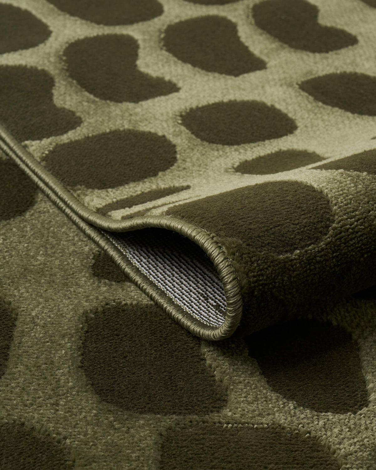 A close-up view of an olive green premium Wilton jacquard rug showing its folded edge, raised pebble-inspired pattern, woven backing, and refined bound finish for elevated living room and bedroom decor.