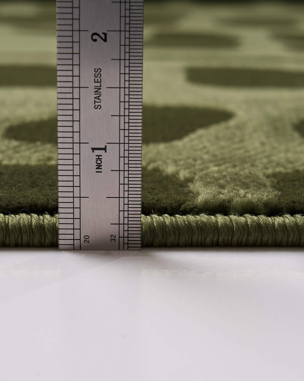 A close-up view of an olive green premium Wilton jacquard rug showing its measured pile height, raised geometric pattern, plush dimensional surface, and refined bound edge for elevated home styling.