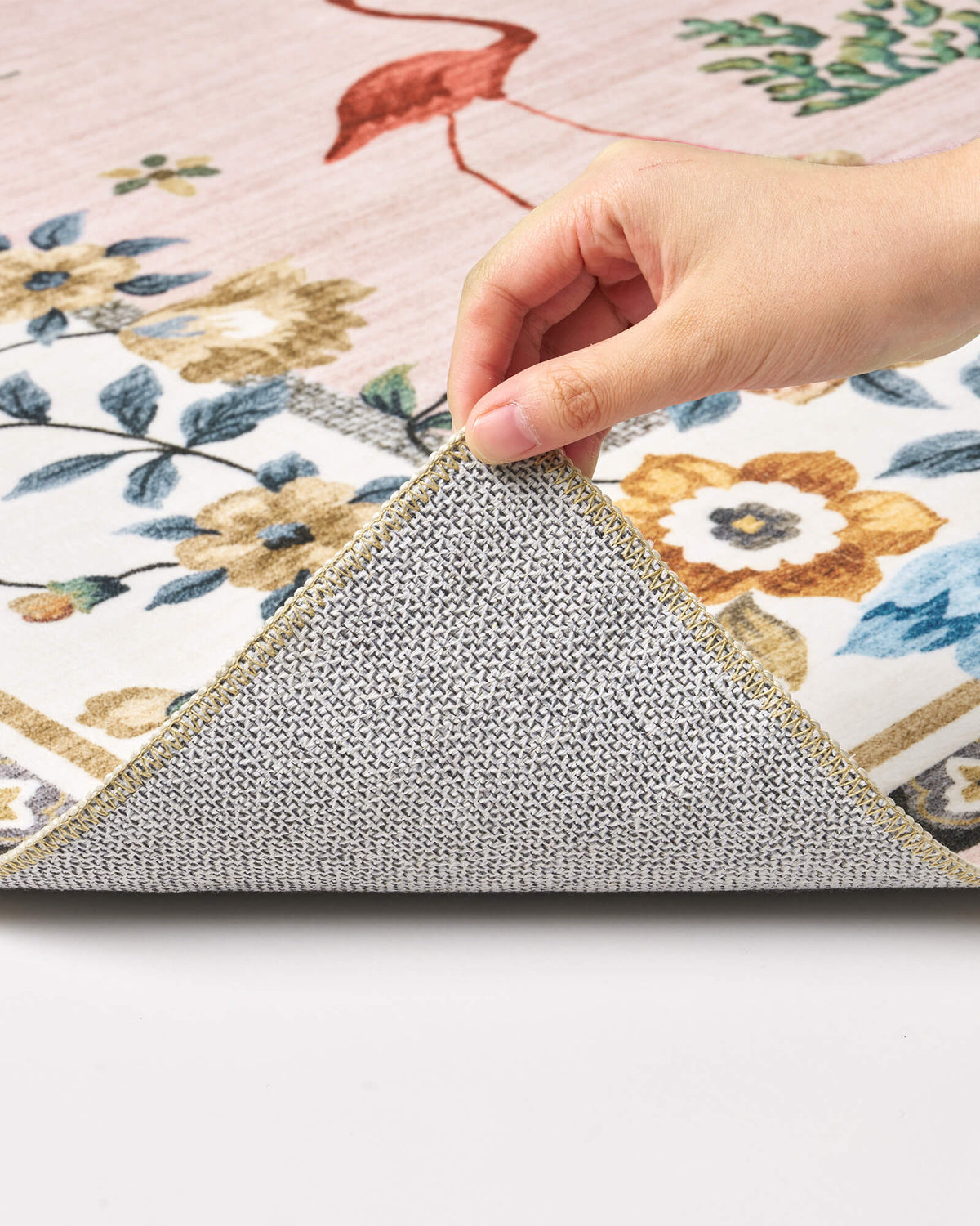 A close-up view of a pink soft kids rug showing its folded corner, gray non-slip backing, flamingo and floral border pattern, and neatly finished edge for safe, cozy nursery, playroom, and children’s bedroom use.