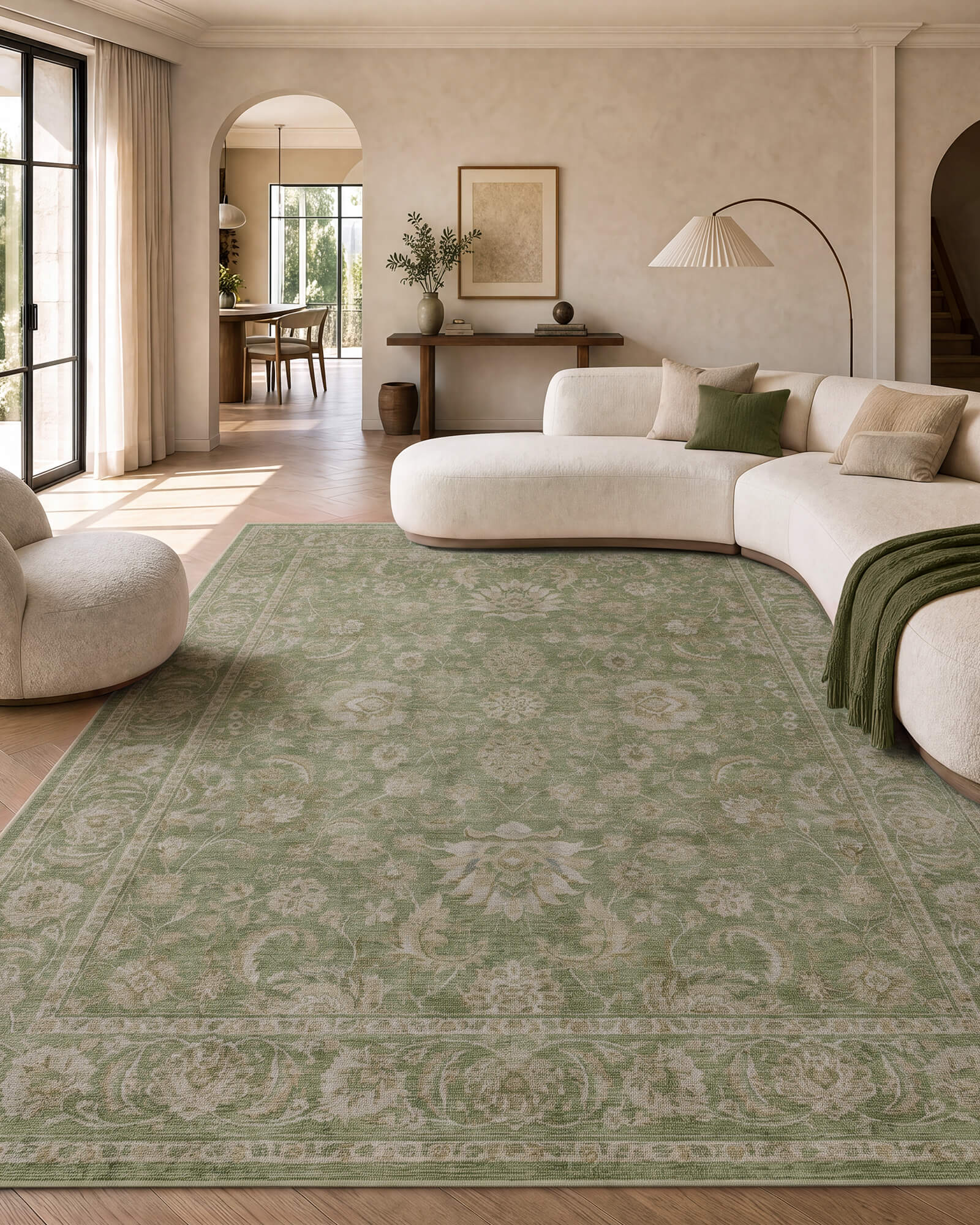 A sage green vintage washable area rug with a soft ornamental border pattern brings calm color to a neutral living room with a curved sectional sofa, warm sunlight, and elegant modern decor.
