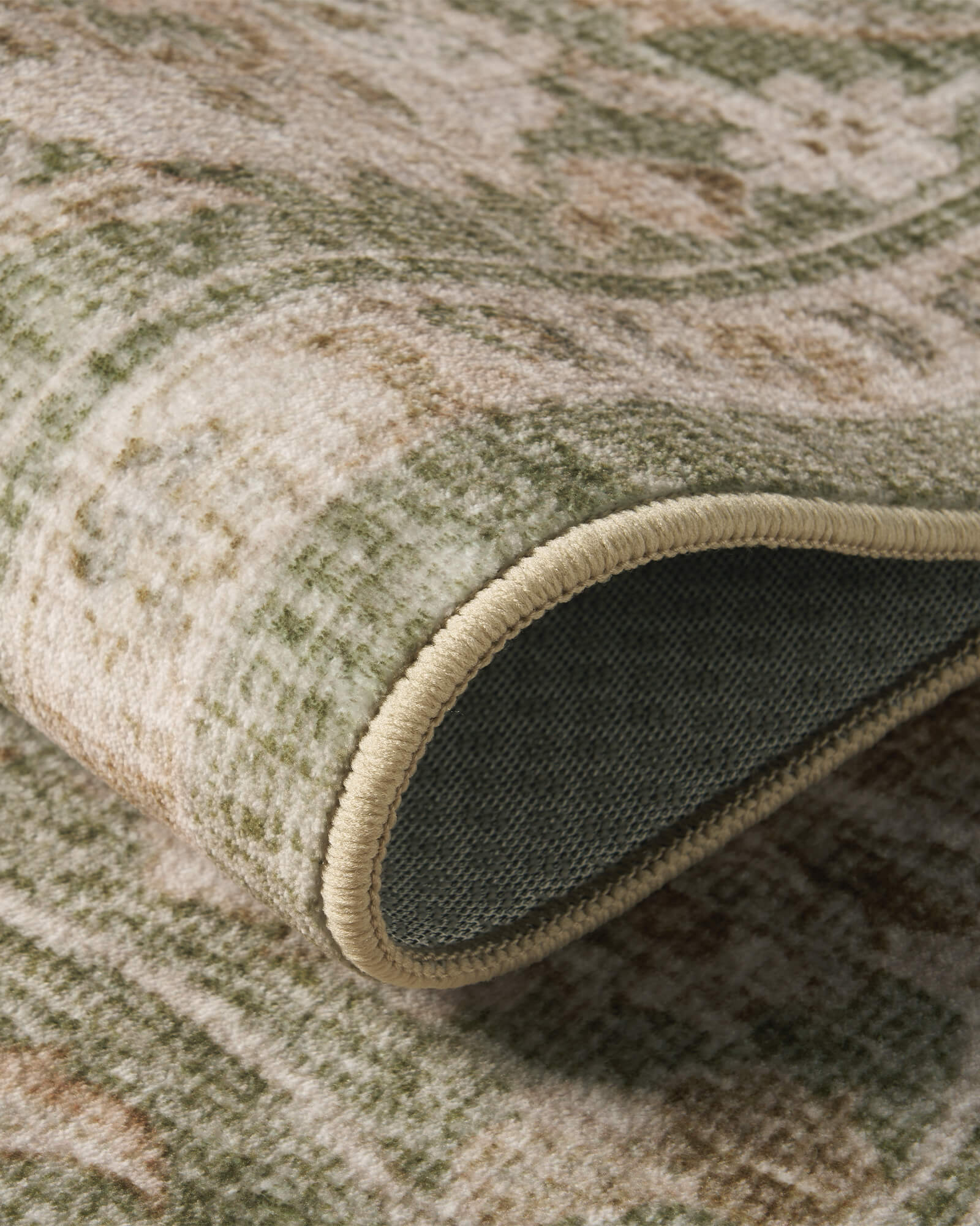 A close-up view of a sage green vintage washable rug showing its curled edge, dark backing, soft ornamental surface, and flexible low-pile construction for comfortable everyday home use.