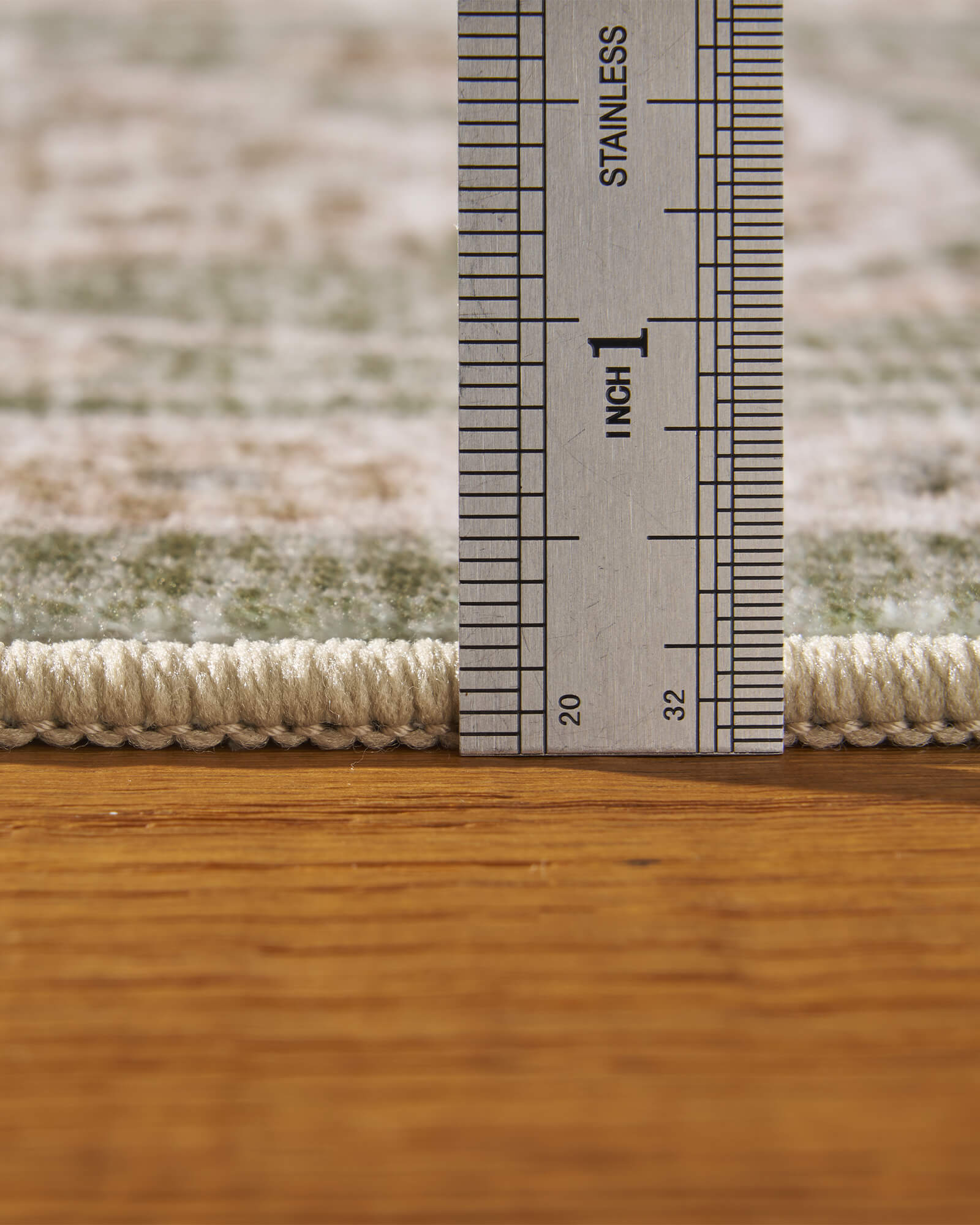 A close-up view of a sage green vintage washable rug showing its ruler-measured pile height, soft low-profile surface, and bound edge for easy placement in bedrooms, living rooms, and family spaces.